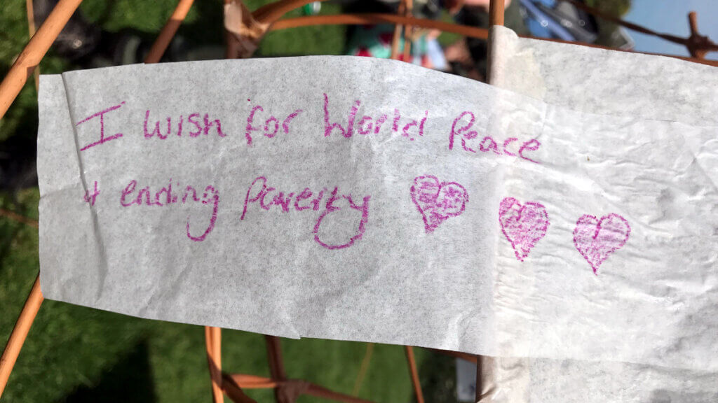 A handwritten wish on a piece of fabric reads ‘I wish for World Peace & ending poverty,’ decorated with three pink hearts, attached to a woven wooden frame in a grassy outdoor setting.