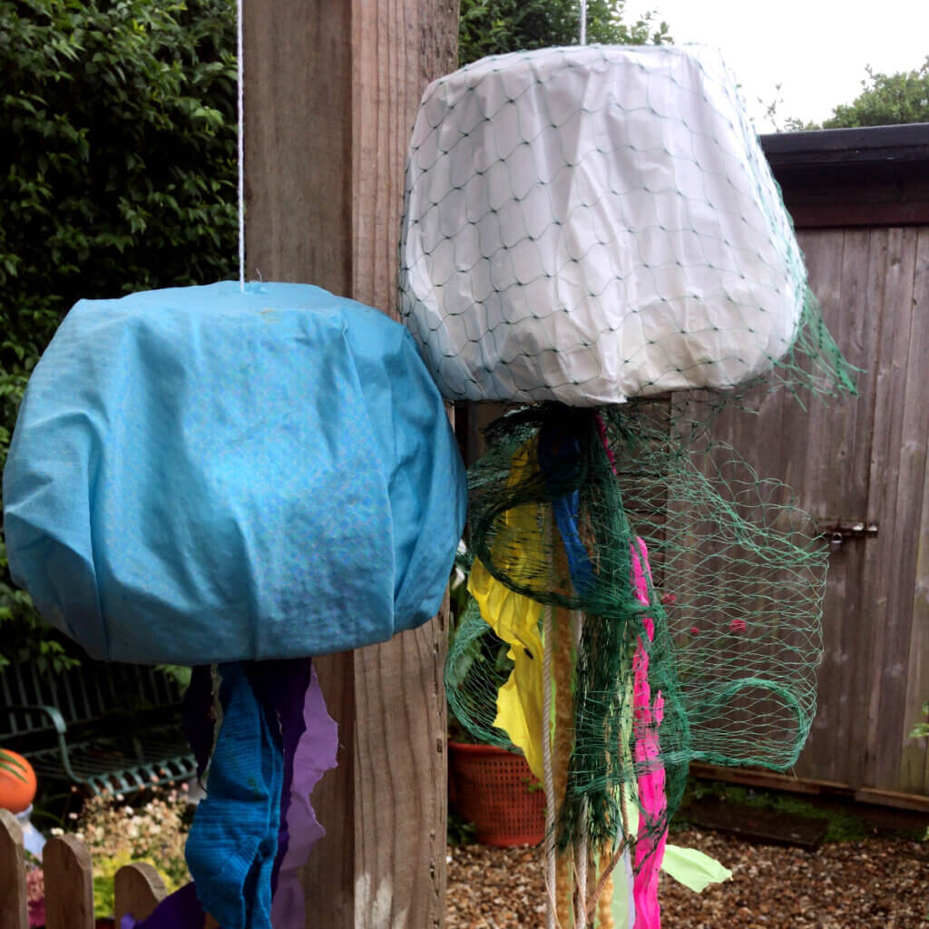 Two jellyfish-shaped lanterns hanging outdoors, one blue and one white with green netting, each with colourful streamers dangling below.