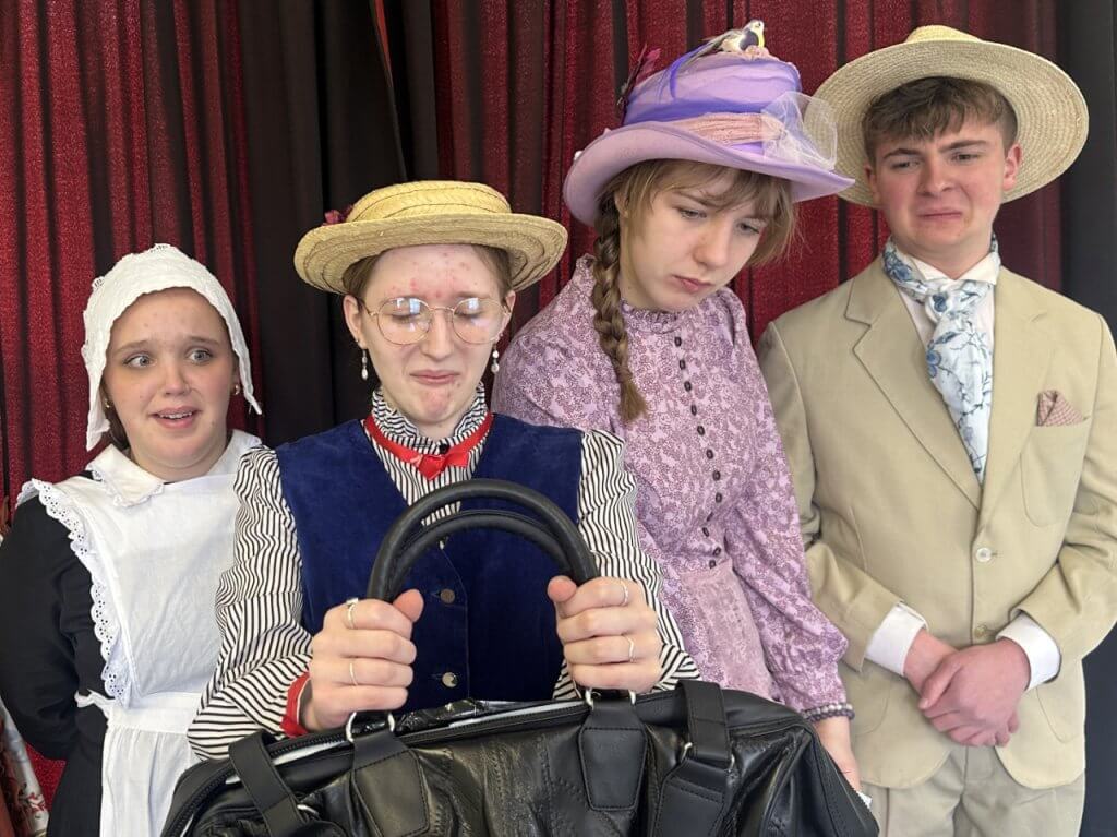 Four young performers in Edwardian costume reacting with comic expressions to a large black handbag, against a red curtain backdrop.