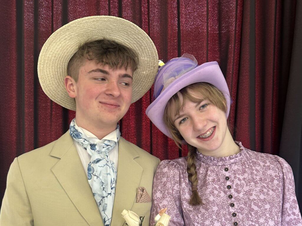 Two young performers in Edwardian costume posing together against a red curtain backdrop.