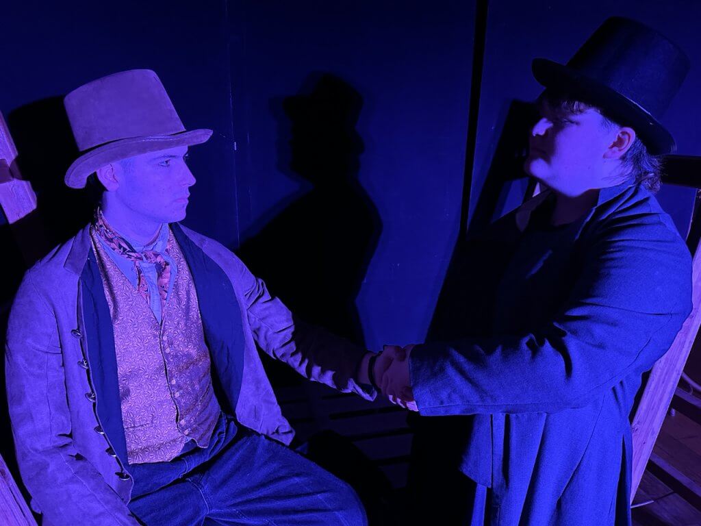 Two young performers in period costume shaking hands dramatically on a darkly lit stage bathed in deep blue light, their shadows cast large on the wall behind them.