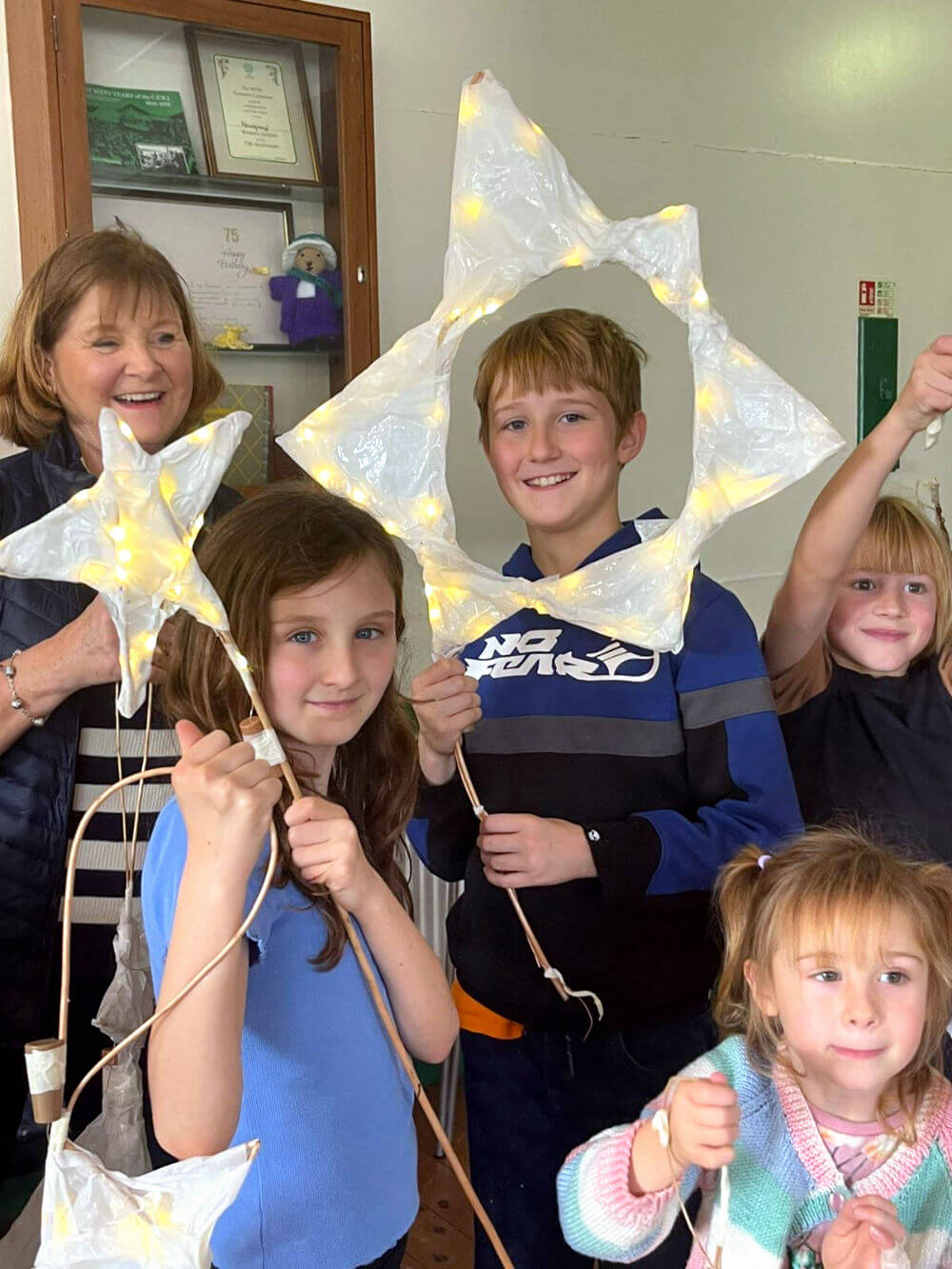 Four children holding up handmade star lanterns made of white tissue-style paper and glowing with warm fairy lights.